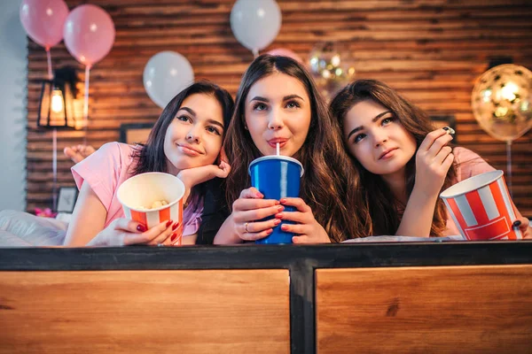 Glada och härliga tonåringar liggande tillsammans på sängen i festlig rum. Flicka i mellersta dricka cola. Andra två modeller har hink med popcorn. — Stockfoto