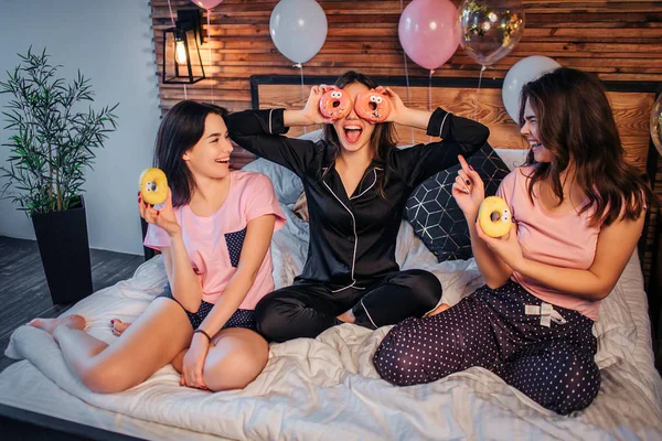 Lustige junge Frau im schwarzen Pyjama sitzen in der Mitte und bedecken die Augen mit rosa Donuts. , lächelt sie. zwei weitere Mädchen haben gelbe Donuts. Sie schauen sie an und lächeln auch. Menschen liegen in festlichem Zimmer auf dem Bett. — Stockfoto