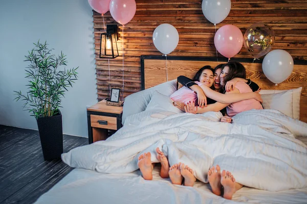 Tres modelos felices yaciendo bajo una manta blanca juntos y abrazando. Sonríen. Las chicas usan pijamas. Los globos están en la cama. Están en la sala de fiestas. . —  Fotos de Stock