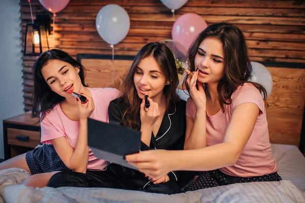 Bild av tre tonåringar gör makeup. De sätta vissa läppstift på läpparna och ser i spegeln. Flickor är koncentrerad. De sitter på sängen i festlig rum. — Stockfoto