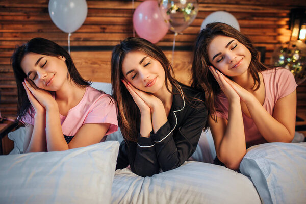Lovely young models sit on bed in room. They wear pajamas. Girls hold heads on hands and keep eyes closed. They are covered with white pillows.
