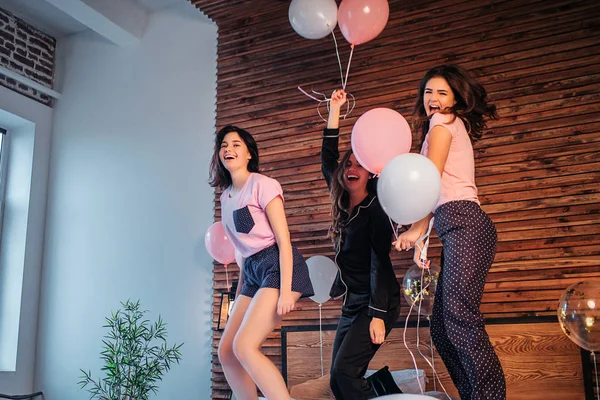 Los adolescentes se divierten en la habitación. Bailan y saltan a la cama. Las mujeres jóvenes sostienen globos y sonríen. Las chicas son felices . — Foto de Stock