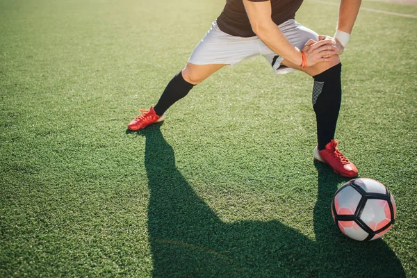 Futbolcu yeşil çimenlerin üzerinde durmak ve bacaklarını uzatmak. O bir-in o eğilir. Dışarıda güzel bir gün. Onun dışında yalan top.
