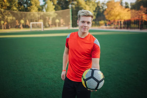 Fröhliche und attraktive junge Spieler halten Ball in der Hand. Er bietet es an. Kerl lächelt. er steht auf dem Rasen. — Stockfoto