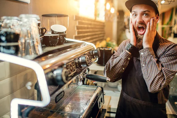 Налякані молодих barista виглядає на камеру. Він тримає обидві руки на щоках. Хлопець стенд на каву-машиною. Він є жаху. — стокове фото