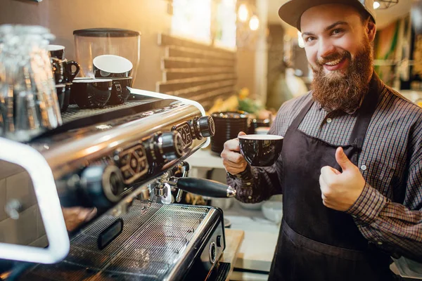 Beau et positif jeune barusta stand dans la cuisine à la machine à café. Il tient un gros pouce et une tasse de café. Guy sourit à la caméra . — Photo