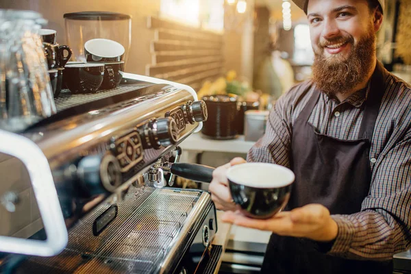 Joyeux jeune homme debout à la machine à café et regarde la caméra. Il montre une grande tasse de café qu'il tient dans ses mains. Guy debout dans la cuisine . — Photo