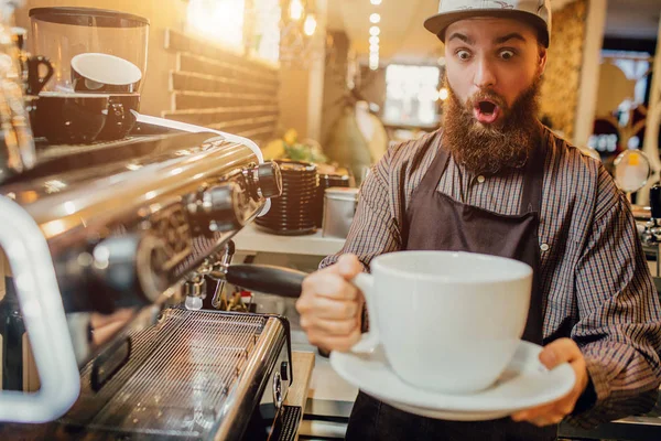 Incroyable jeune homme barbu tenir énorme tasse de café blanc dans les mains. Il le regarde avec excitation. Guy stand à la machine à café dans la cuisine . — Photo