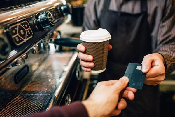 Photo de barista se tenir à la machine à café et tenir une tasse de café. Il prend la carte d'un autre homme pour payer le café. . — Photo