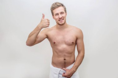 Pozitif genç adam kameraya bakıyor. O göstermek büyük başparmak ve tutun havlu el arounf kalçalarda. Adam mutlu. Beyaz arka plan üzerinde izole.