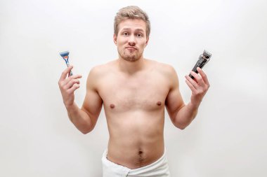 Şaşkın genç oğlan razers ve tıraş makinesi elinde. Kameraya bakıyor. Gömleksiz biri. Beyaz arka plan üzerinde izole.
