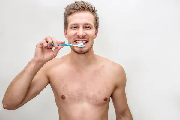 Добре побудований і без сорочки молодий чоловік чистить зуби зубною щіткою і зубною пастою. Він дивиться на камеру. Ізольовані на білому тлі . — стокове фото