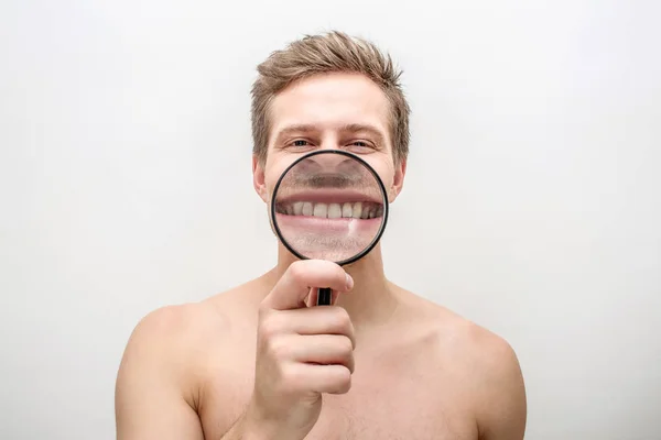 Happy young man showing his teeth through magnifying glass. He looks straight. Isolated on white background. — Stock Photo, Image
