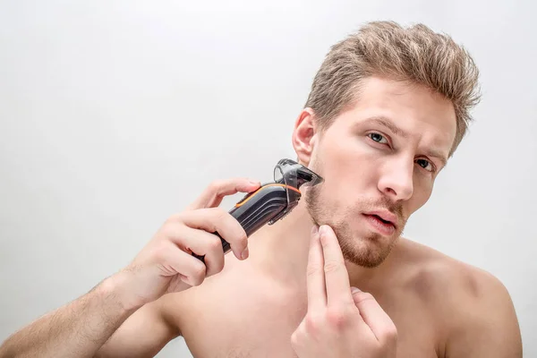 Allvarlig ung man rakning. Han använder rakning maskin. Han arbetar med kindområdet. Isolerad på vit bakgrund. — Stockfoto