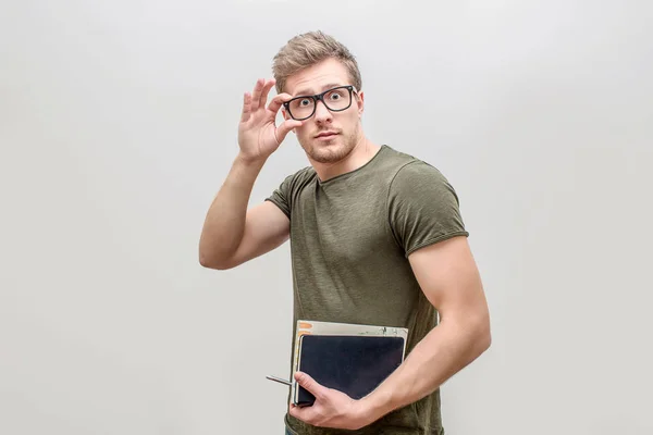 Well-build and strong young man posing on camera. He looks straight and hold hand on glasses. Guy has books in other hand. Isolated on white background. — Stock Photo, Image