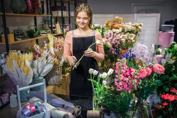 Joven florista alegre parada en la tienda de flores. Ella sostiene el tulipán blanco y corta parte del tallo. Ella sonríe . — Foto de Stock