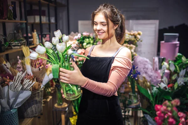 Joven alegre sostiene jarrón de flores de vidrio con tulipanes blancos. Ella sonríe. Floristería stand en floristería . — Foto de Stock