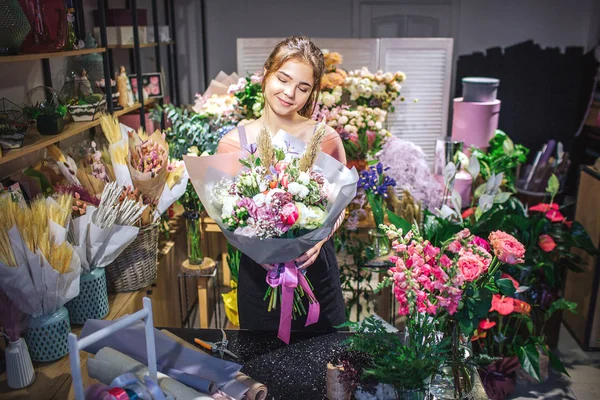 Schöne und attraktive Floristin ist unter bunten Blumen. Sie hält einen Blumenstrauß in den Händen und betrachtet ihn. junge Frau lächelt. — Stockfoto