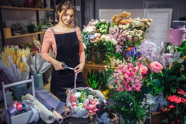 Nice oung woman stand in room full of flowers and plants. She look down and cut violet ribbon. It it tight to bouquet on table.