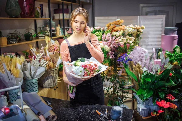 Ocupado florista joven tiene ramo en la mano. Ella habla por teléfono. Mujer joven de pie en la habitación llena de flores y plantas . — Foto de Stock