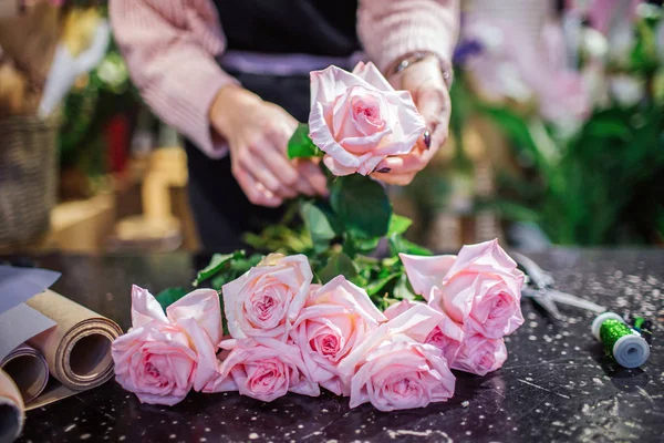Cierre de rosas rosa claro beautfiul acostado en la mesa. Floristería tiene una flor en la mano. Rolld de papel y bobina con hilo verde están en la mesa . — Foto de Stock
