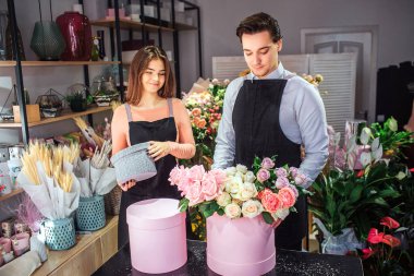 Neşeli genç çiçekçi masada hazır. Adam gül pembe kutuya koy. Genç kadın küçük gri kutu tutun. Onlar gülümseme. Genç adam işine konsantre olması.