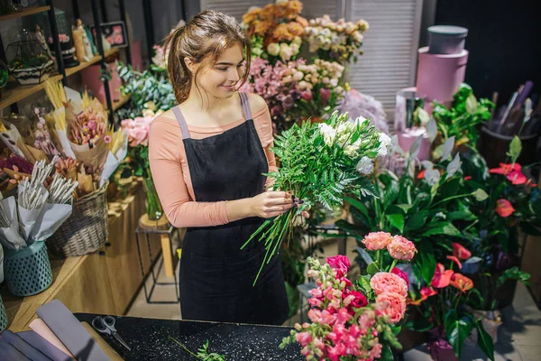 Jovem alegre ficar e combinar flores. Ela faz bouquet. Um sorriso florista. Ela está na sala cheia de flores e plantas . — Fotografia de Stock