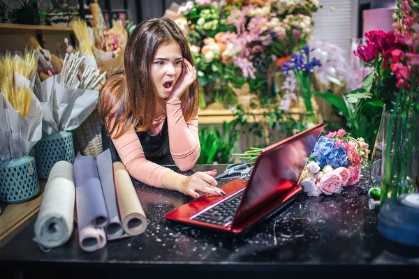 年轻的女花店在桌子上的照片。她看着笔记本电脑, 纳闷。纸卷在桌子上。许多花和植物都在花店的后面. — 图库照片
