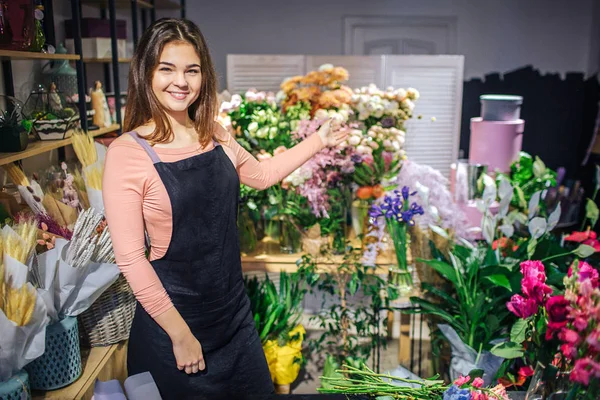 Jovencita alegre posando en cámara. Sonríe y muestra plantas y flores con la mano. Mujer joven usar delantal negro . — Foto de Stock