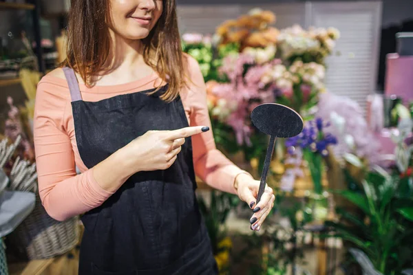 Geschnittene Ansicht von oyung weibliche Floristin Punkt auf schwarze Holzsache in der Hand. Sie steht vor Blumen und Pflanzen. junge Frau trägt schwarze Schürze. — Stockfoto