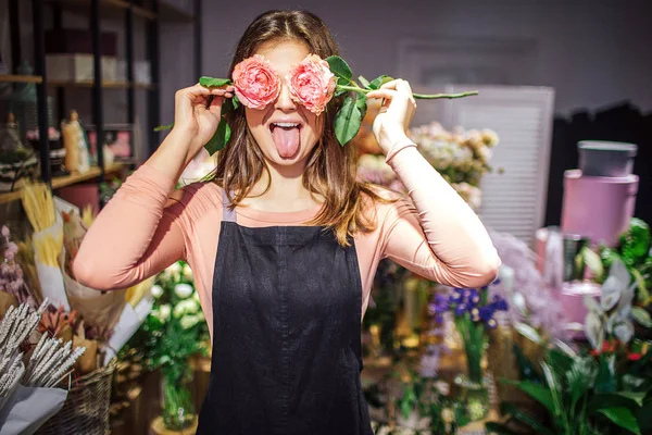 Lustige und positive junge Floristin bedeckt Augen mit Rosen und zeigt Zunge. Sie hat Spaß. Dahinter stehen Pflanzen und Blumen in Vasen. — Stockfoto