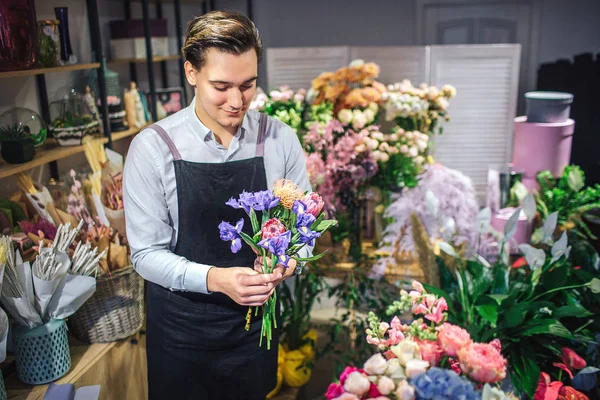 Joven florista alegre del varón sostiene el ramo colorido en las manos. Él lo mira. Diferentes plantas y flores están detrás de él. Guy de pie dentro . — Foto de Stock