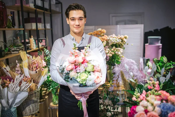 Hermoso joven florista masculino de pie y mirar en cámara. Tiene un ramo de flores en las manos. El tipo está dentro. Él alegre . — Foto de Stock