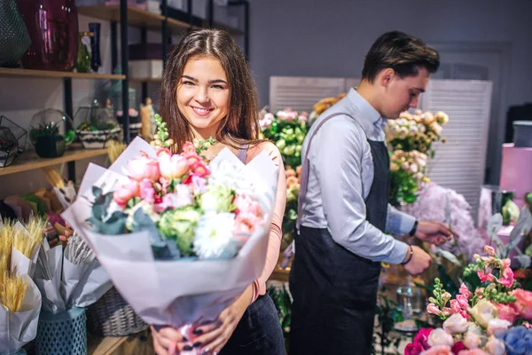 Fröhliche junge Floristin blickt in die Kamera und lächelt. Sie hält einen Blumenstrauß in den Händen. Hinter ihr stehen männliche Floristen. sie sind drinnen. — Stockfoto