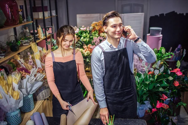 Young woman stand at table and cut piece of paper rolls. Guy stand behind her and talk on phone. He smile. They are in room full of flowers and plants.