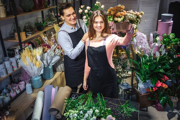 Floristerías femeninas y masculinas jóvenes están en una habitación llena de flores y plantas. Sujeta a Hone. Se toman selfie. La gente sonríe y posa . — Foto de Stock