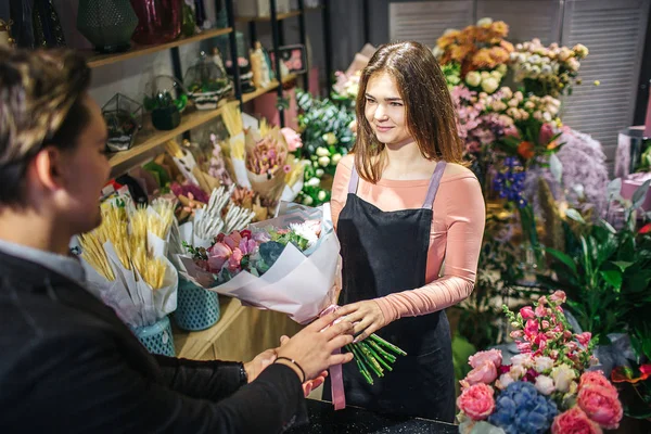 Junge feamel Florist halten Blumenstrauß und schauen Kunden an. sie lächelt. Mann in Jacke schaut sie an und greift nach Blumen. sie stehen drinnen. — Stockfoto