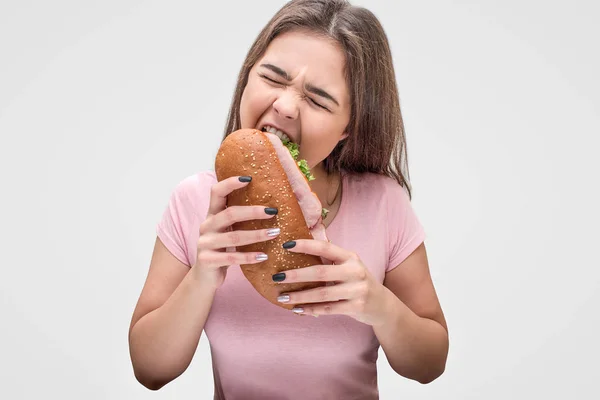 Młoda kobieta pożreć sandwich. Ona trzymać go w ręce i oczy zamknięte. Na białym tle na szarym tle. — Zdjęcie stockowe