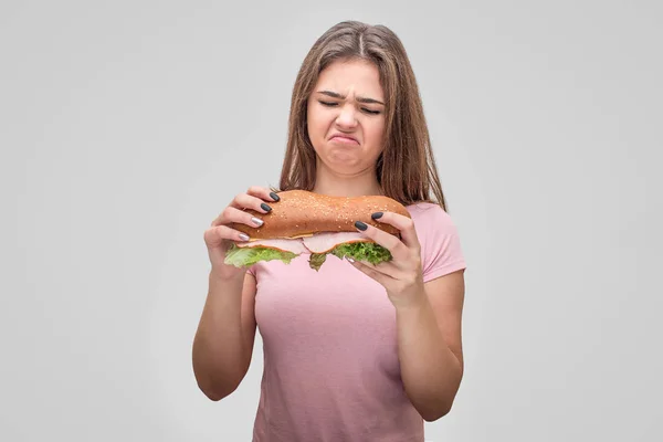 Ongelukkige jonge vrouw houd hamburger en staar ernaar. Ze krimpt. Geïsoleerd op grijs bakground. — Stockfoto