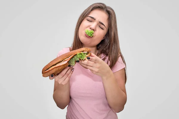 Jonge vrouw heeft stuk groene sla in mond. Ze heeft lekkere hamburger. Model genieten. Geïsoleerd op een grijze achtergrond. — Stockfoto