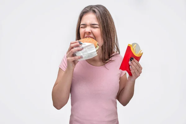 Wanita muda lapar menggigit sepotong burger. Dia memegang kentang goreng di tangan lain. Terisolasi pada latar belakang putih . — Stok Foto