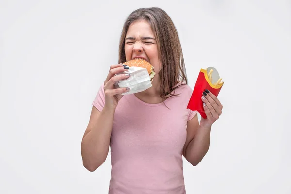 Hungry young woman bite piece of burger. She hold fried potatoes in another hand. Isolated on white background. — Stock Photo, Image