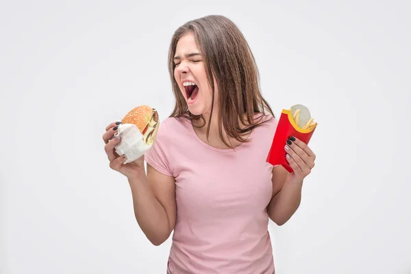 Junge Frau schreit bei geöffneter Tür. Sie hält Burger und Kartoffeln in der Hand. isoliert auf grauem Hintergrund. — Stockfoto