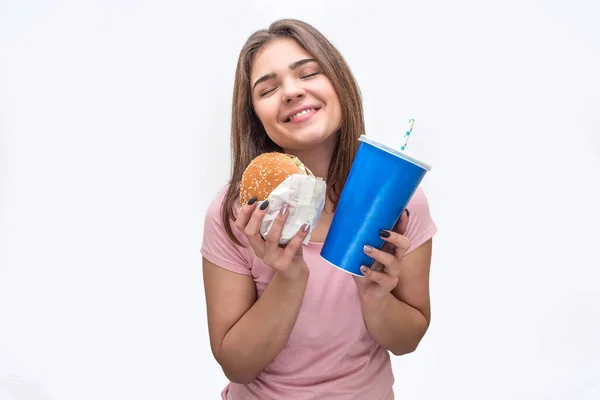 Vrolijke en gelukkige jonge vrouw houd hamburger en kopje coca-cola in handen. Ze ziet er geweldig. Geïsoleerd op witte achtergrond. — Stockfoto