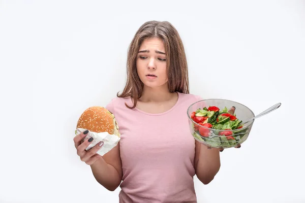 Young woman hold burger and glass bowl of salad. She looks at junk food. Model doubts. Isolated on grey background. — Stock Photo, Image