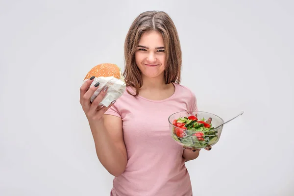 Jovens mulheres agradáveis e positivas olham para a câmera. Ela segura hambúrguer mais alto do que tigela com salada. Isolado sobre fundo cinzento . — Fotografia de Stock