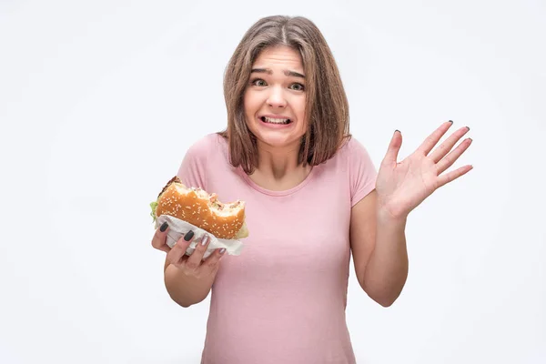 Korkmuş ve hayal kırıklığına uğramış genç kadın tutuşsun ısırıldı burger. O palm gösterir. Gri arka plan üzerinde izole. — Stok fotoğraf