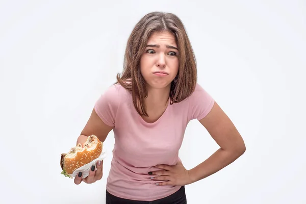 Foto di giovane donna rutto. Tiene una mano sullo stomaco e un'altra con hamburger. La modella sta male. Isolato su sfondo bianco . — Foto Stock