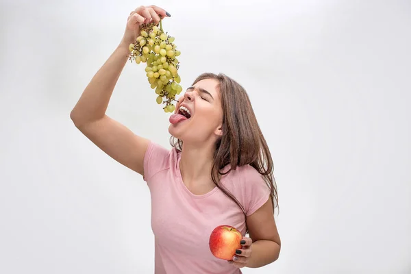 Фото молодой женщины, держащей виноград над открытым ртом. Она доходит до него языком. Яблоко в другой руке. Изолированный на сером фоне . — стоковое фото