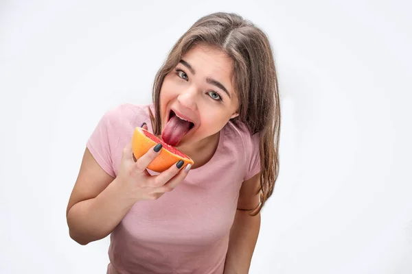 Grappige jonge vrouw likt stukje grapefruit. Ze ziet er rechte. Geïsoleerd op witte achtergrond. — Stockfoto